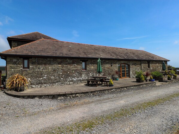 A stone building with a tiled roof, a gravel driveway, and outdoor seating with umbrellas.