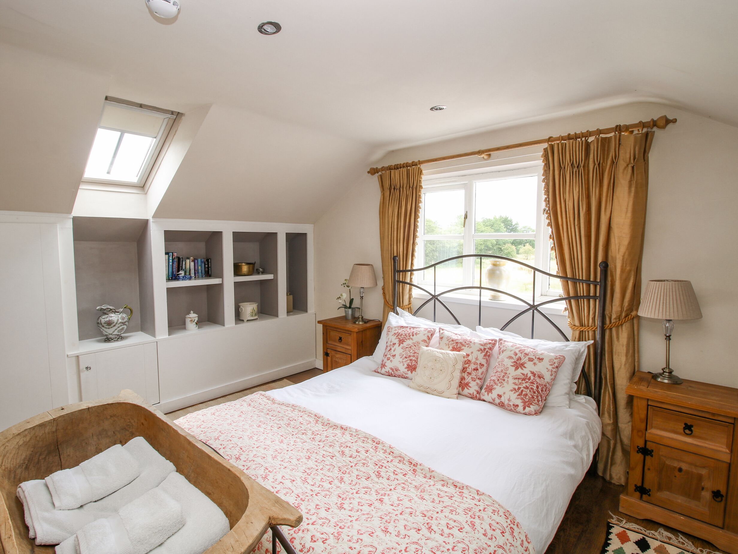 A bedroom with a bed, a wooden nightstand, a bedside table, a window with curtains, a skylight, and built-in shelving.