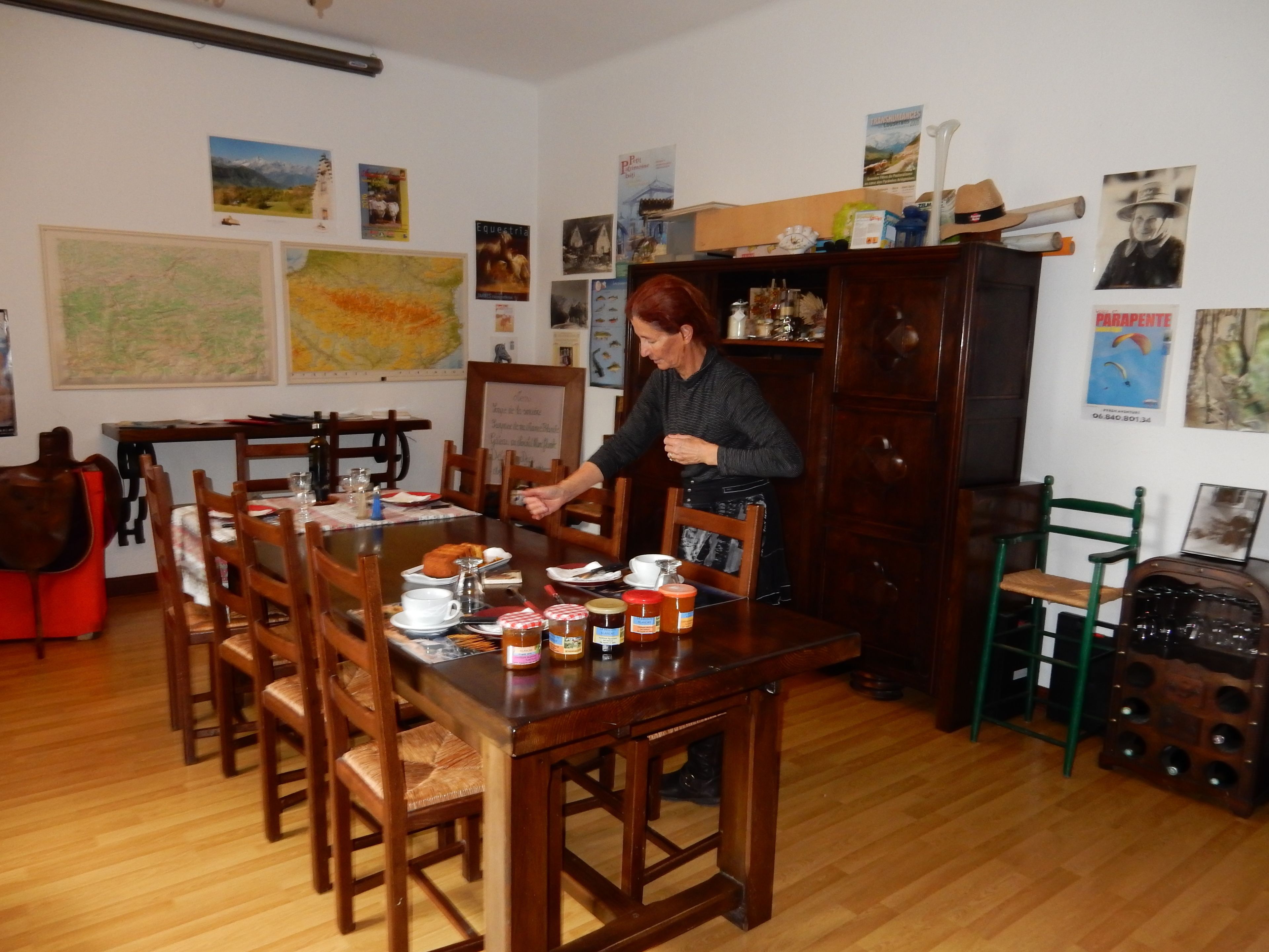 A dining area with a wooden table set for four, surrounded by wooden chairs. There are various items on the table, including plates, cups, and jars. The walls are decorated with framed pictures and posters.