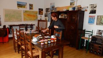 A dining area with a wooden table set for four, surrounded by wooden chairs. There are various items on the table, including plates, cups, and jars. The walls are decorated with framed pictures and posters.