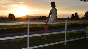 A person standing by a white fence in a rural setting with a sunset in the background.