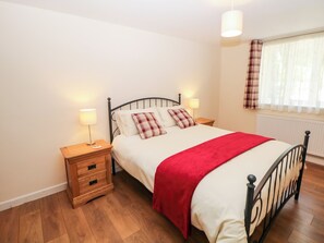 A bedroom with a bed, two bedside tables, a nightstand, a red throw blanket, a lamp, a window with curtains, and a radiator.