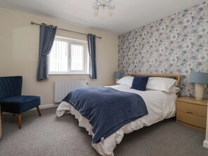 A bedroom with a bed, a chair, a nightstand, a lamp, a window with curtains, and a radiator. - The Milking Parlour (York)