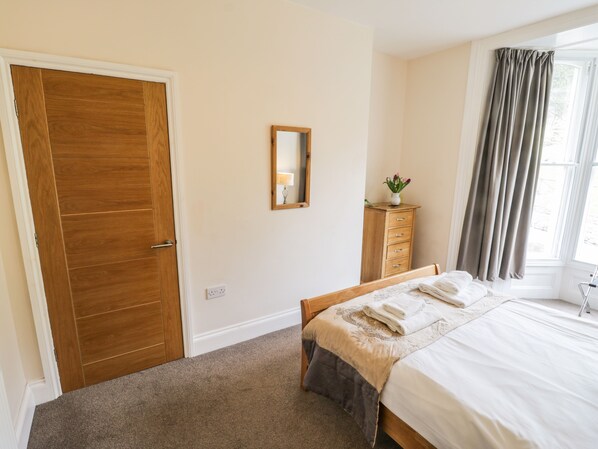 A bedroom with a wooden door, a bedside table with a lamp, a bed with a white comforter, and a window with curtains.