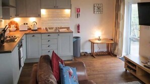 A compact kitchen with a built-in oven, a coffee maker, and a toaster. There is a small table with a lamp and a fire extinguisher on the wall. A brown sofa with colorful cushions is in the living area, and a TV is mounted on the wall. - The Old Corn Store (Keighley)