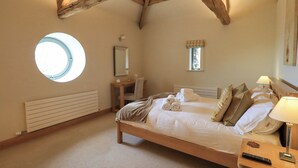 A bedroom with a wooden ceiling, a round skylight, a bed with neatly folded towels, a bedside table with a lamp, and a small desk with a mirror.
