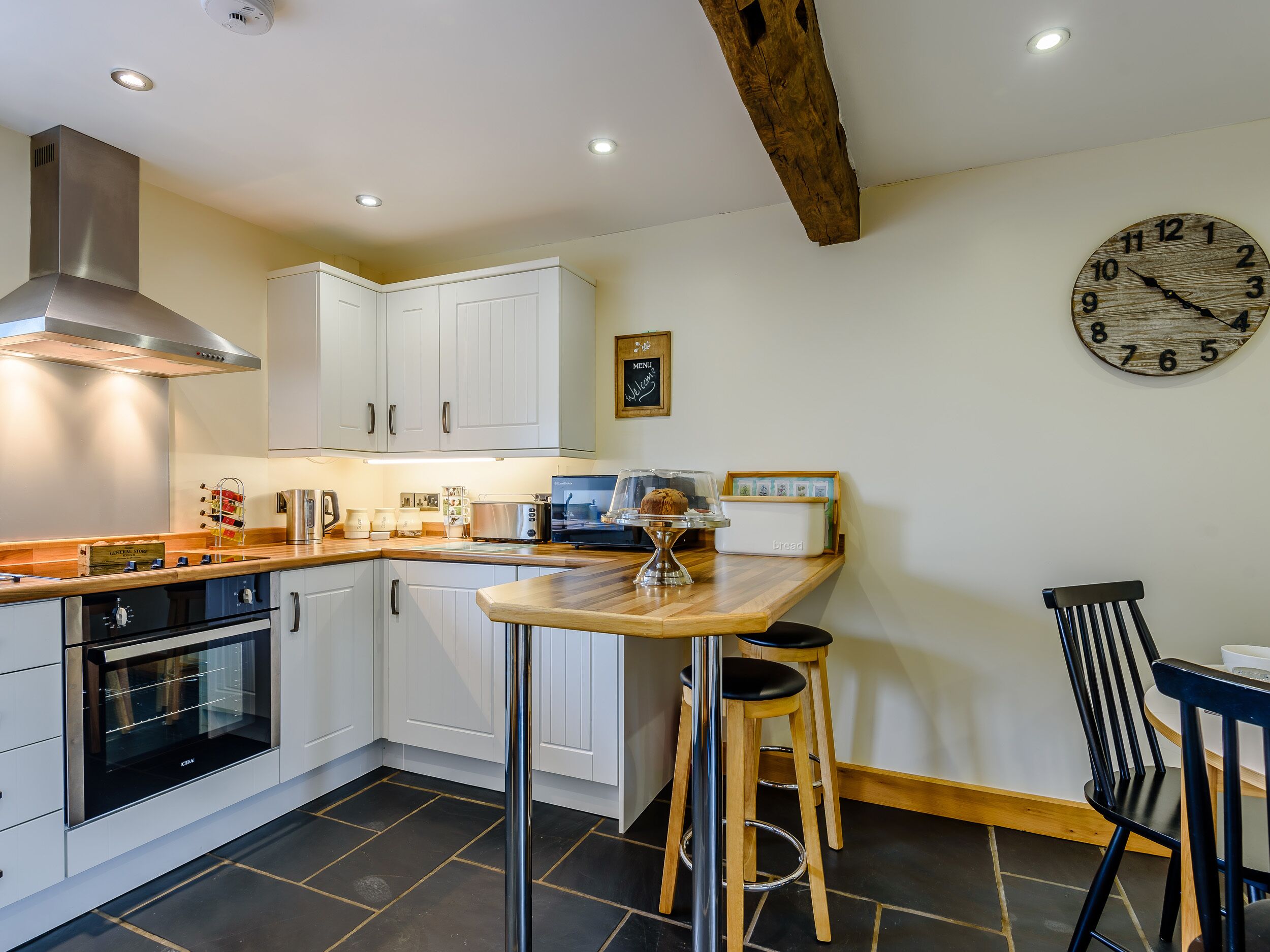 A modern kitchen with white cabinets, a stainless steel range hood, a built-in oven, a coffee machine, and a bread box.
