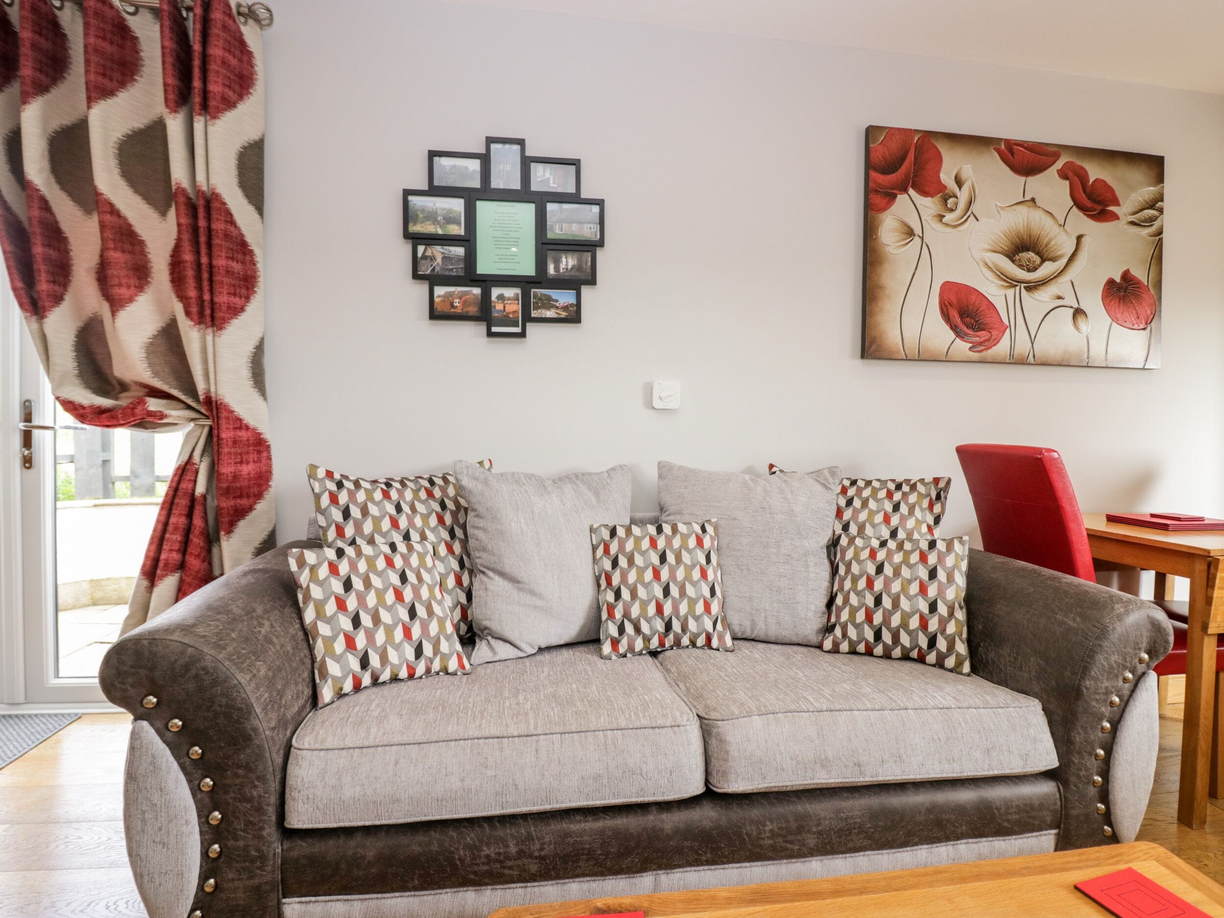 A living room with a grey sofa, patterned cushions, a red chair, a wooden table, and a wall with framed pictures and a floral painting.