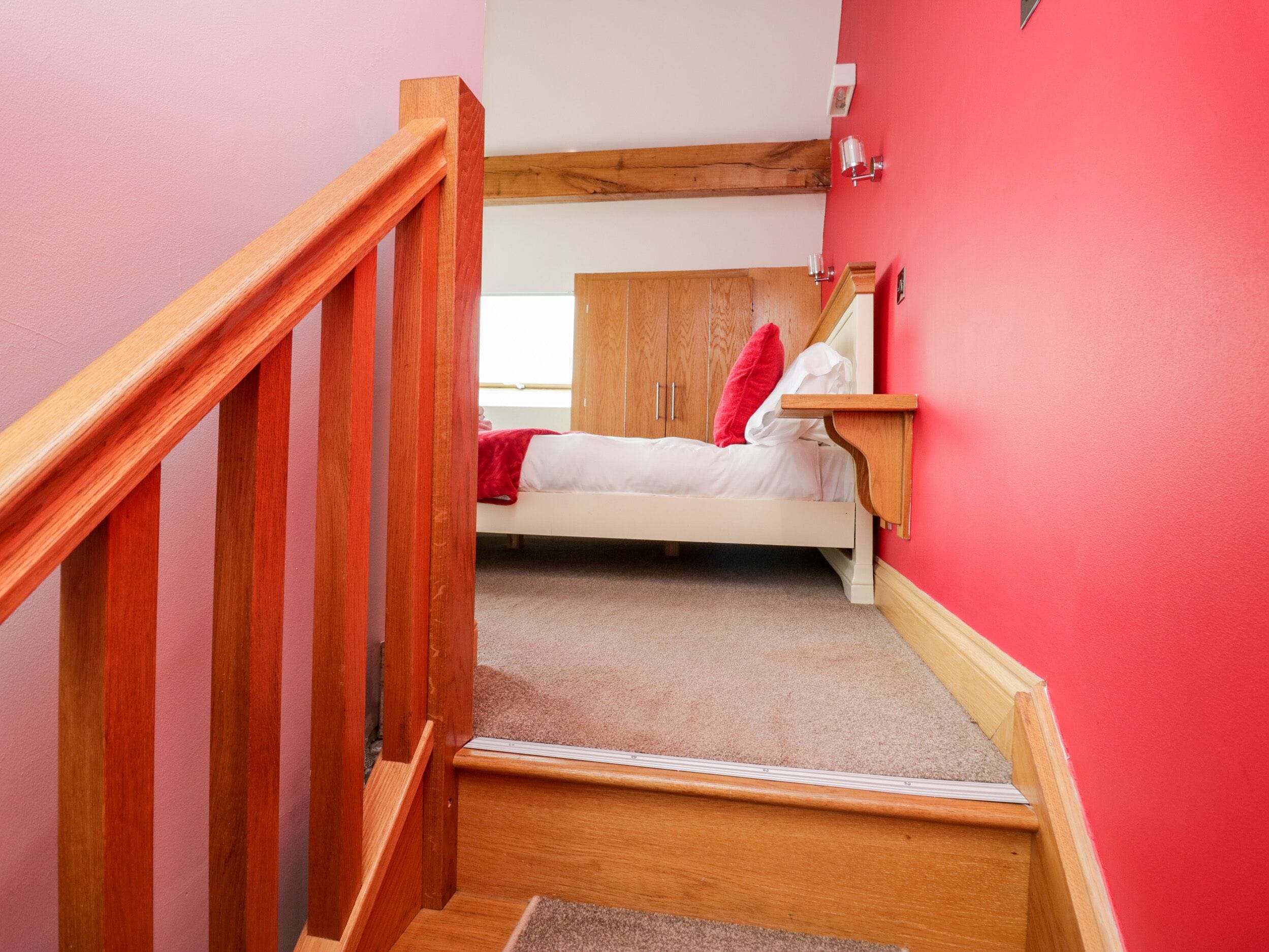 Une chambre avec un escalier en bois, un lit avec une literie blanche et un oreiller rouge, et une tête de lit en bois.