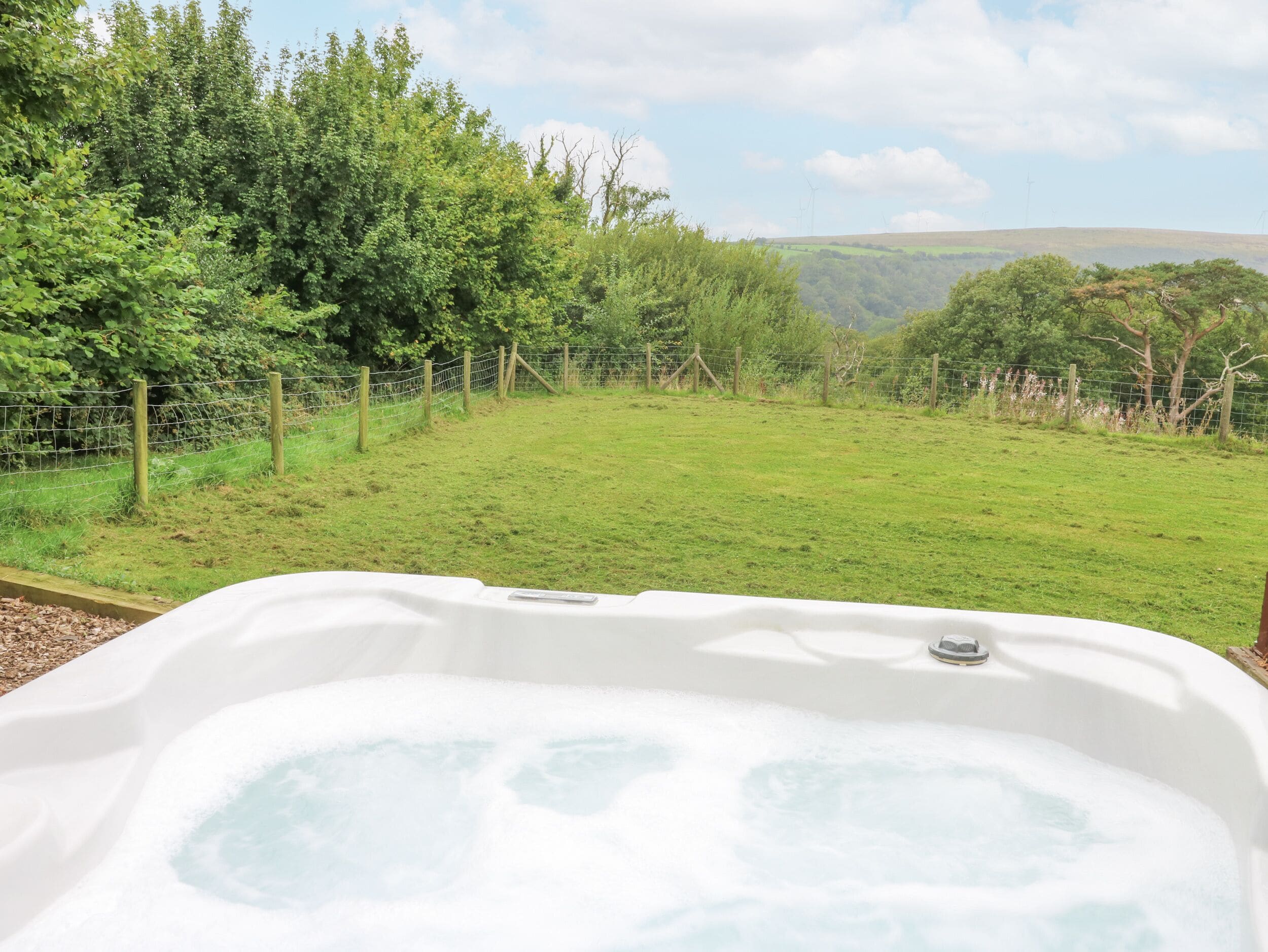 A hot tub in a lush green garden with a view of hills and wind turbines in the distance.