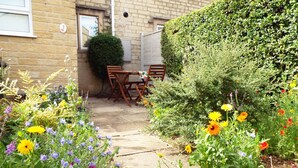 A garden pathway leading to a wooden table and chairs, surrounded by various flowering plants and a hedge.