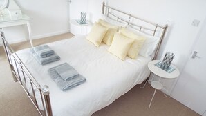 A neatly made bed with white linens and multiple pillows, a small bedside table with a glass object, and a white nightstand with a similar item.