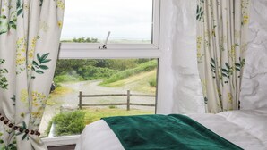 A bedroom with a window overlooking a rural landscape, curtains with floral patterns, and a green throw on the bed. - Hendy Cottage (Llanbedr)
