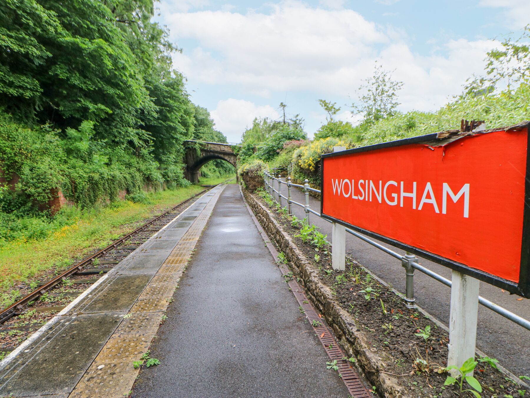 Un andén de tren con un letrero rojo que dice "WOLSSINGHAM" y un puente de piedra al fondo.