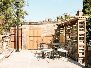 A stone patio with a wooden gate, a metal table with chairs, and a wooden pergola. - Rose Cottage (High Peak)