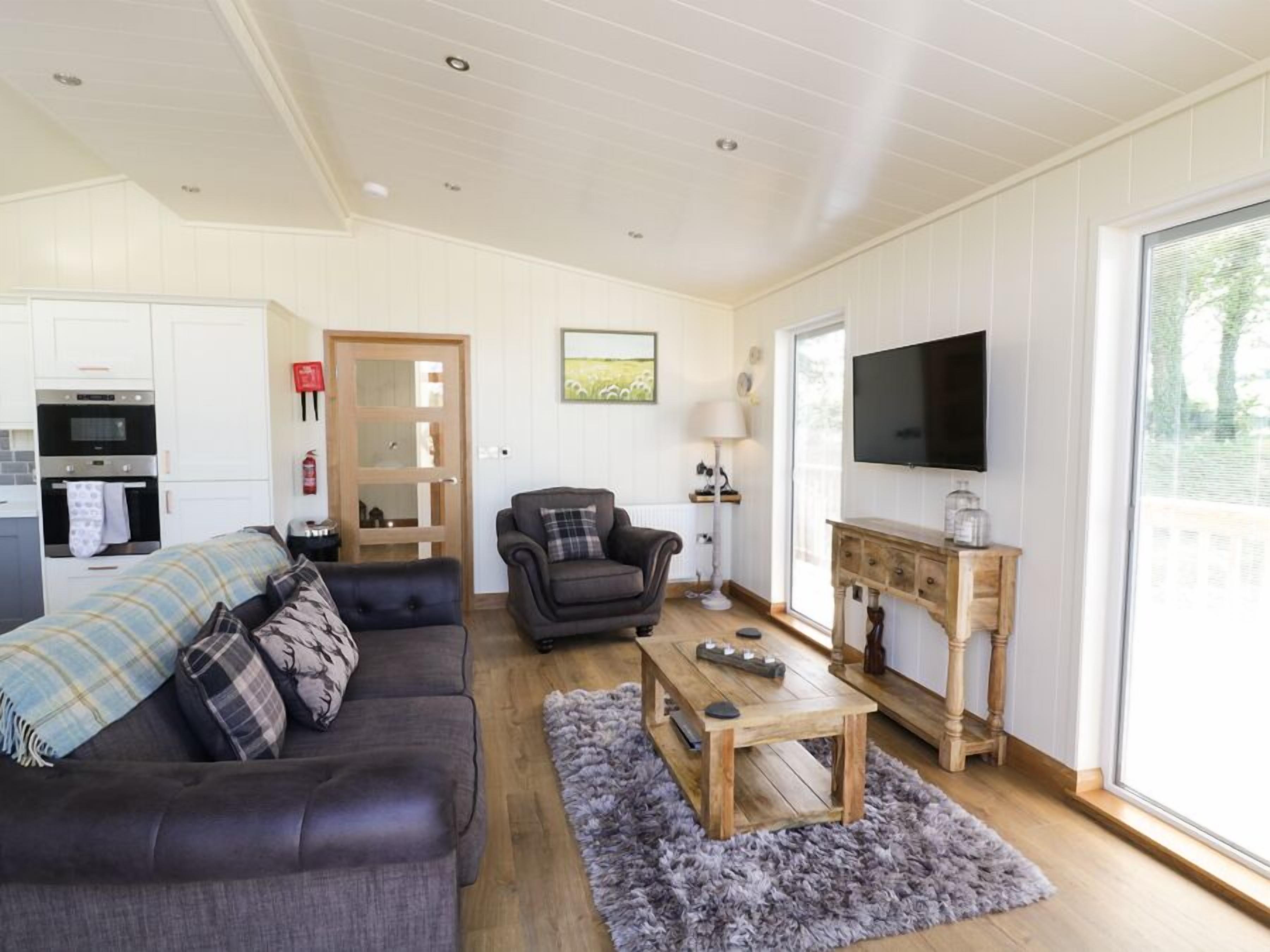 A living room with a sofa, armchair, coffee table, and a TV. There's a kitchen area with an oven and a door.