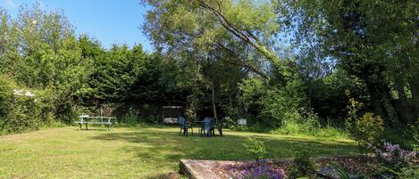 Un espace extérieur verdoyant, avec une table de pique-nique et des chaises, entouré d’arbres et d’un jardin agrémenté de fleurs violettes.