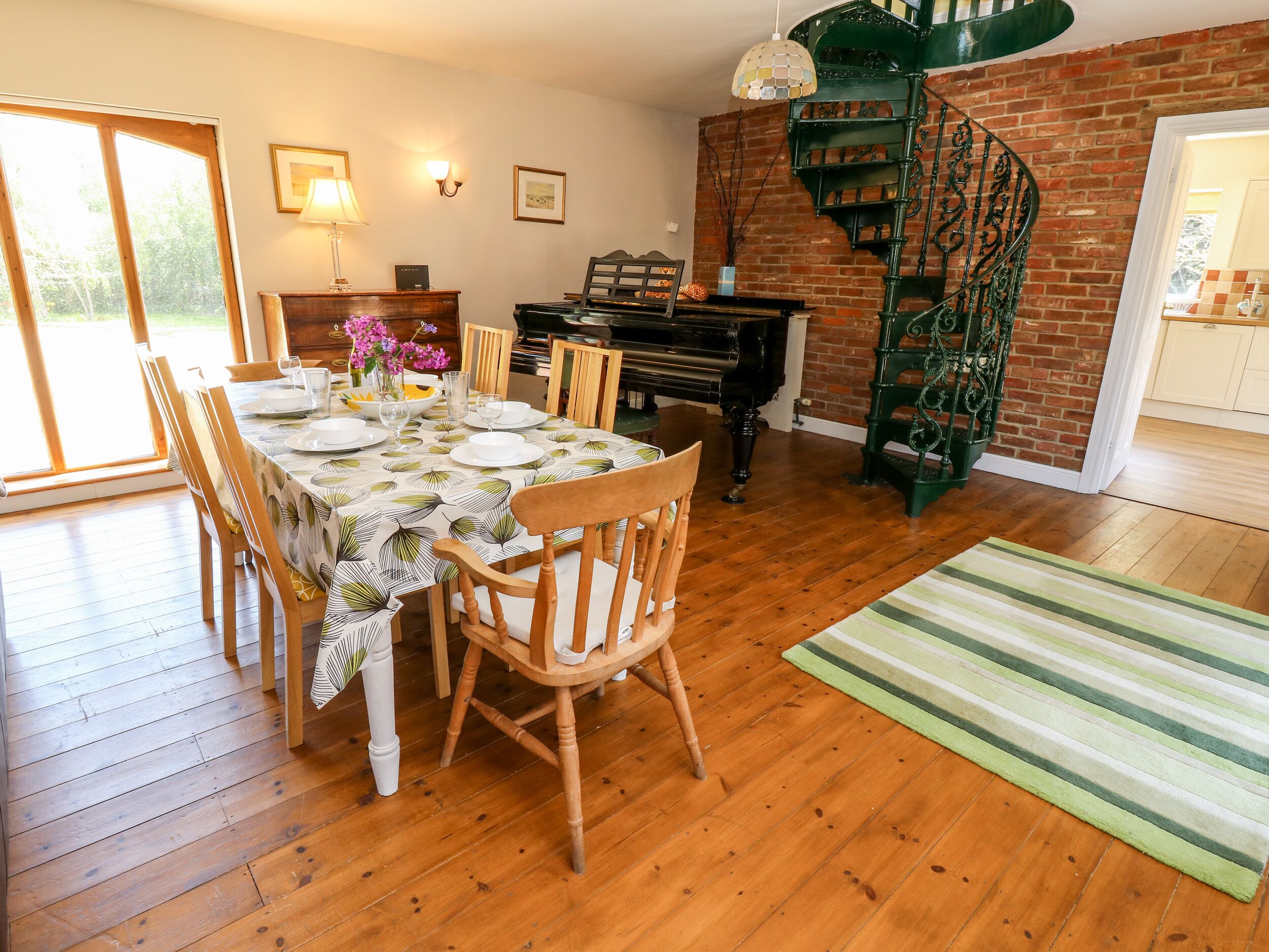 Un coin repas avec un piano, des chaises en bois, une table dressée pour quatre personnes et un escalier en colimaçon adossé à un mur de briques.