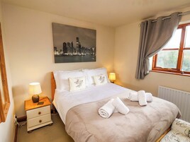 Una cama bien hecha con sábanas blancas y toallas enrolladas, una mesita de noche con lámpara, un cuadro de un paisaje urbano en la pared y una ventana con cortinas.
