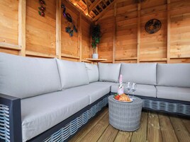 Interior de una cabaña de madera con un sofá gris, una mesita de mimbre con una botella y vasos, y decoración en la pared.