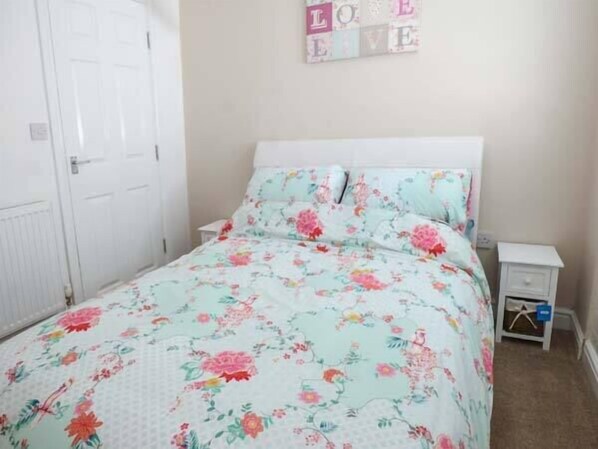 A bedroom with a floral bedspread, a white headboard, a bedside table with a lamp, and a wall decoration that reads "LOVE LIVE". - Flat 3 (Bridlington)
