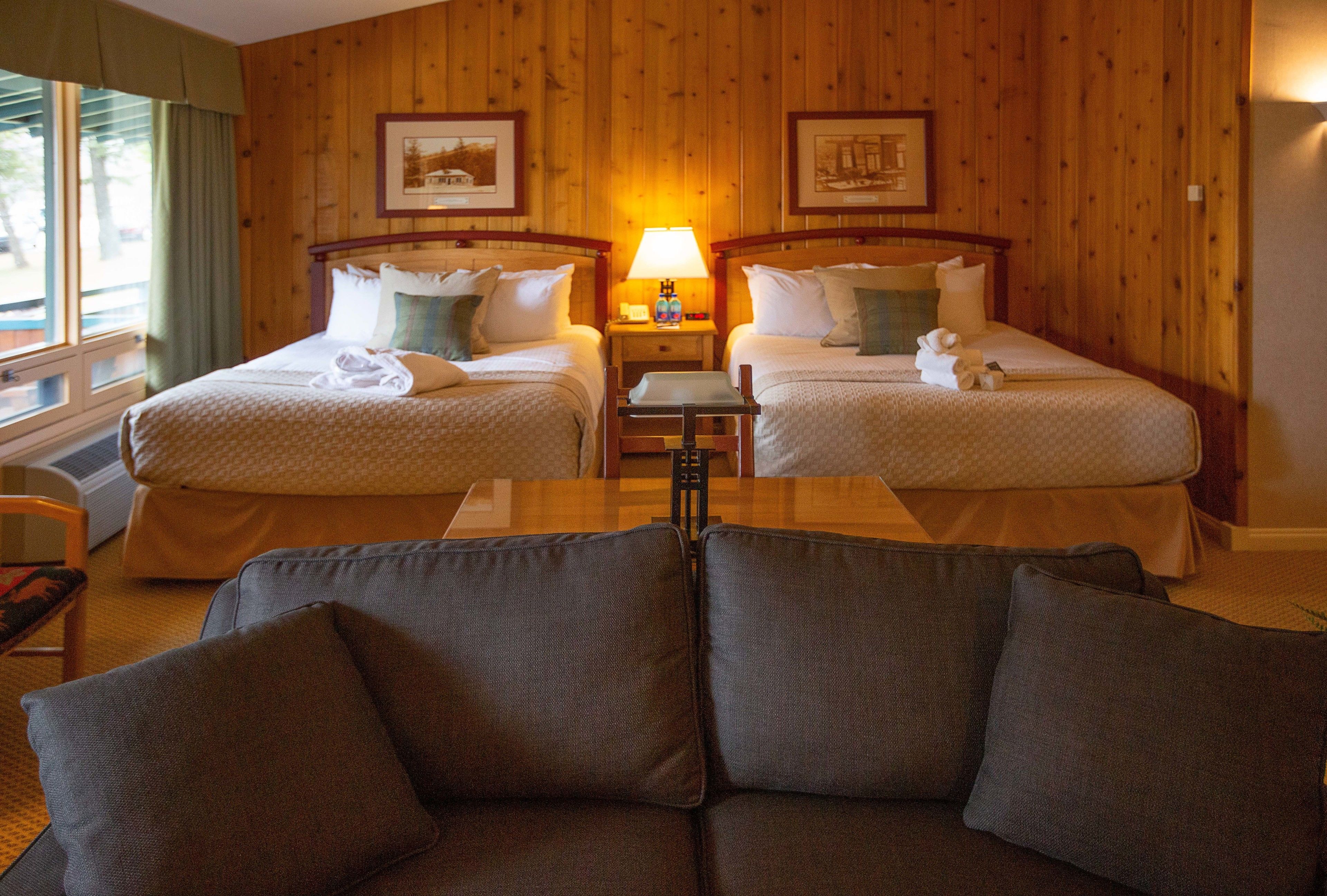A hotel room with two beds, a wooden floor, a wooden headboard, a nightstand with a lamp, and a sofa.