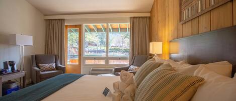 A hotel room with a large bed, two armchairs, a desk with a lamp, and a view of the outdoors through a window.