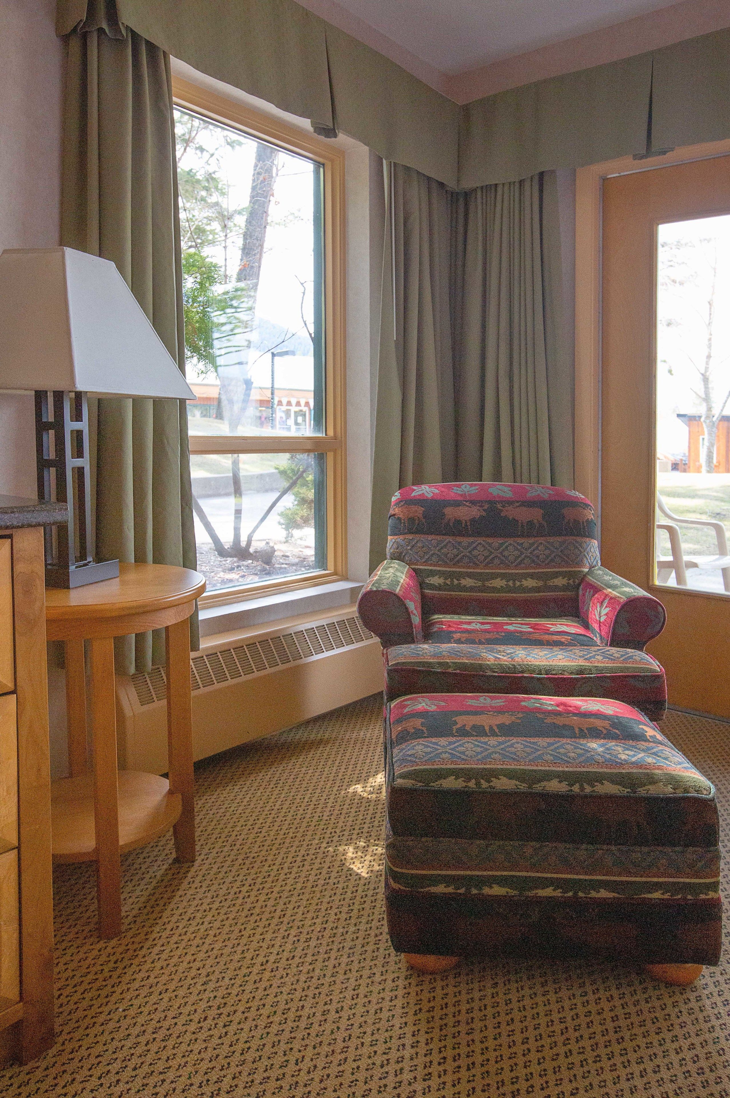 A corner room with a patterned armchair and ottoman, a small wooden table with a lamp, and a window with curtains.