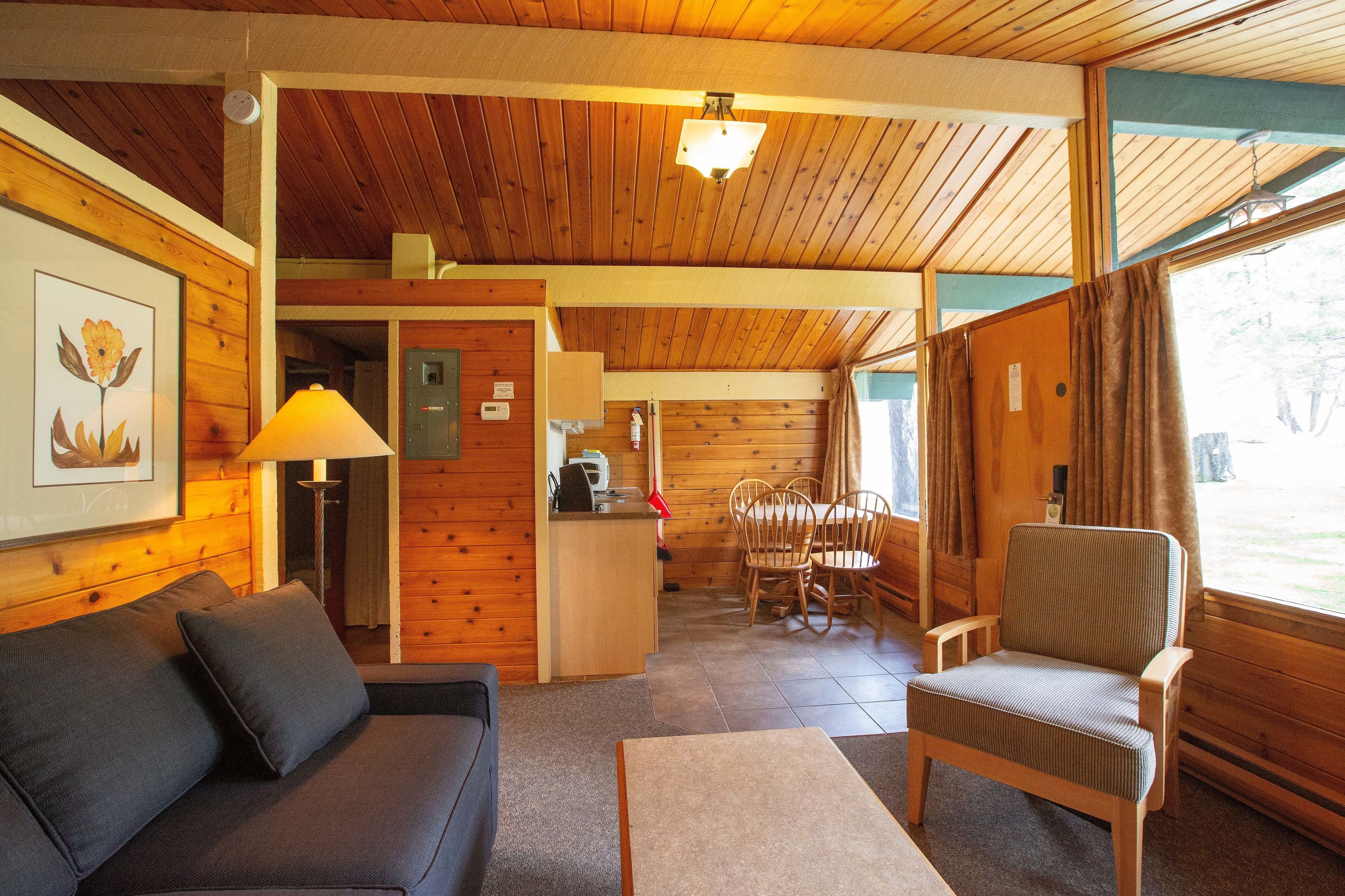 Interior de una cabaña de madera con sofá, sillón, mesa de comedor y zona de cocina.