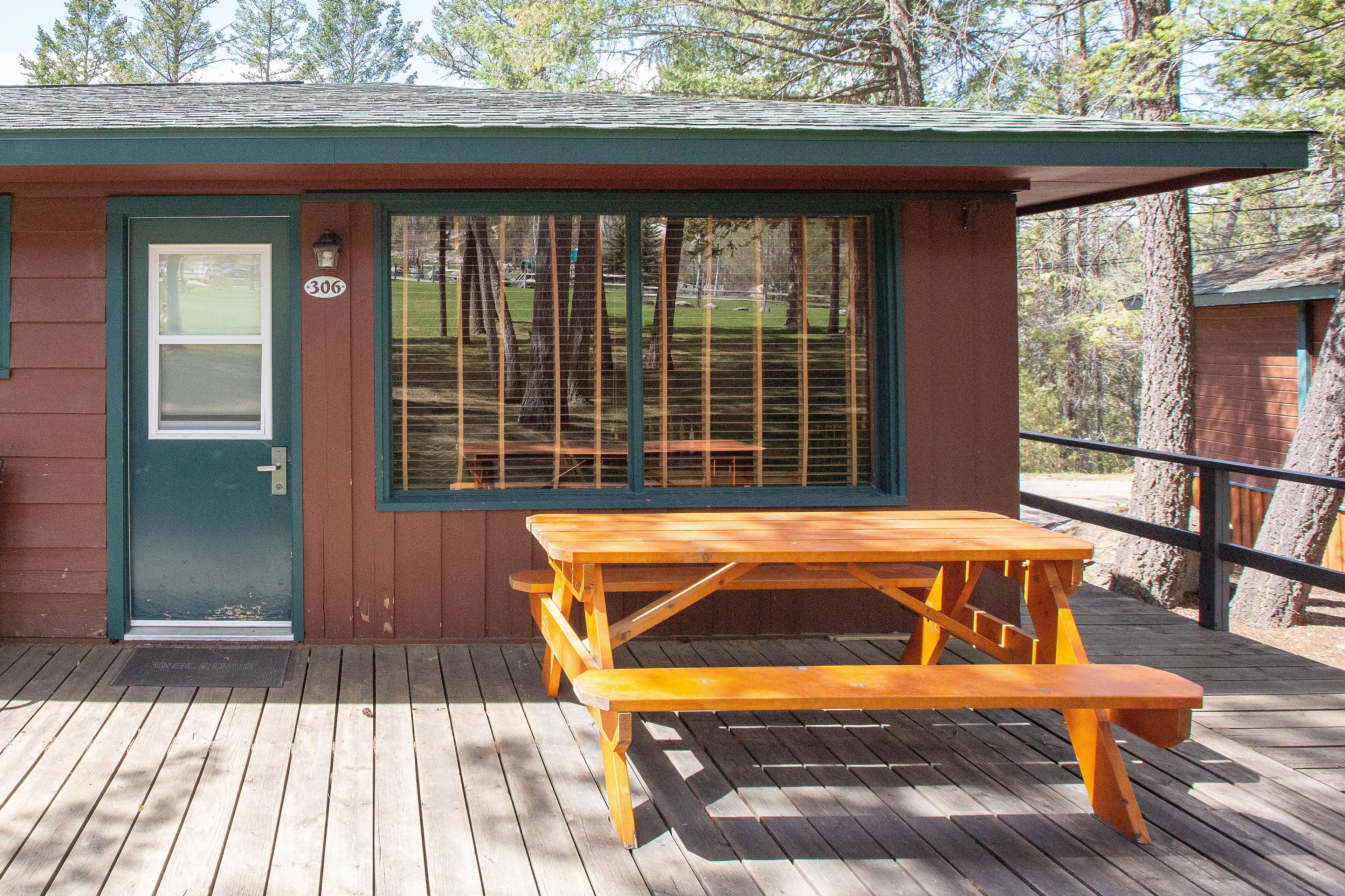 A wooden picnic table and bench on a deck, with a green door and window, and a number 306 sign on the door.