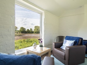 A room with a large window, a brown leather sofa, a small wooden table with a vase and a book, and a view of a green landscape.