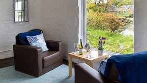 A room with a brown leather sofa, a small wooden table with a bottle of wine and glasses, and a view of a garden and houses through a large window. - Bay View (Belford)