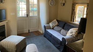 Une pièce confortable avec un canapé, un pouf et un téléviseur. On y trouve une porte avec une fenêtre, une horloge au mur et une lampe.