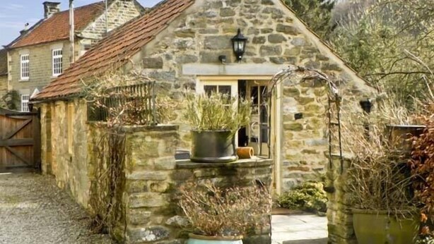 Une petite maison en pierre avec un toit en pente, une porte en bois et des plantes en pot devant l’entrée.