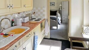 Une petite cuisine équipée d’un évier, d’une cuisinière et de placards. On aperçoit un lit à travers une porte ouverte.