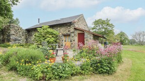Ein Steinhäuschen mit roter Tür, umgeben von einem Garten mit verschiedenen Blumen und Pflanzen.