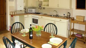 Una cocina con armarios blancos, microondas, estufa y lavaplatos. Hay una mesa de comedor con sillas y una botella de vino.