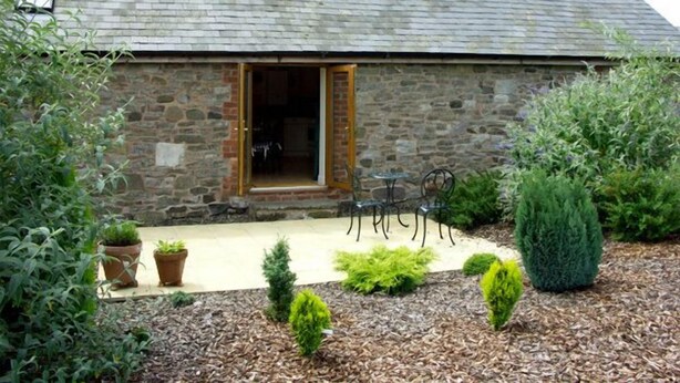 Un edificio de piedra con puerta de madera, un pequeño patio con mesa y sillas, y diversas plantas en macetas.