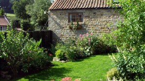 Une maison en pierre avec un toit en tuiles, entourée d’un jardin bien entretenu où poussent différentes plantes et fleurs.