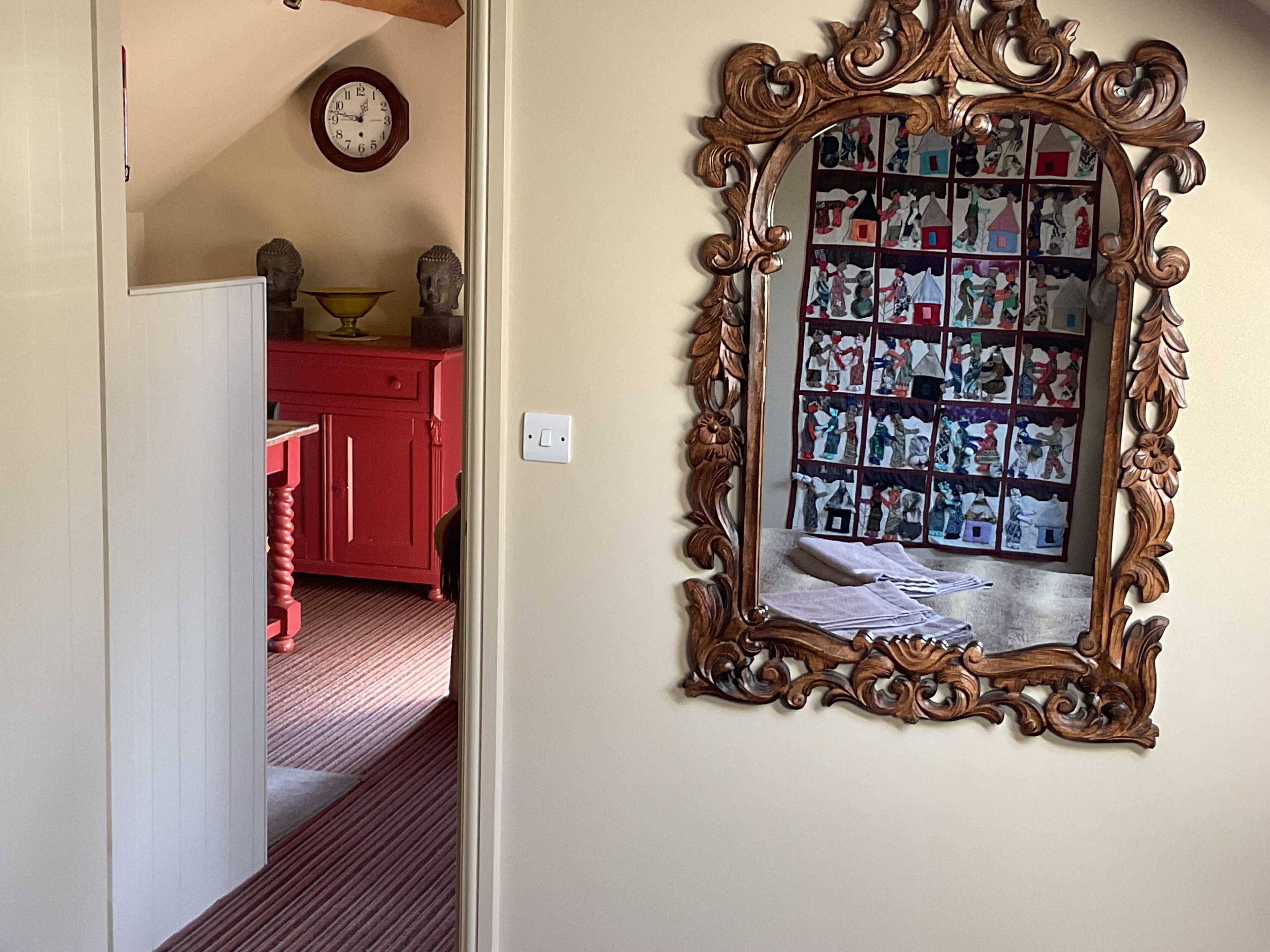 A hallway with a wooden-framed mirror reflecting a room with a red cabinet and a clock on the wall.