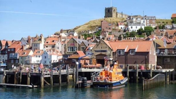 Une ville côtière dotée de bâtiments historiques, d’une tour imposante et d’un quai où est amarré un bateau.