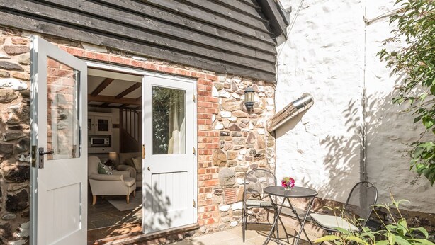 A stone house with a white door, a small patio with a table and chairs, and a garden with yellow flowers. - The Old Workshop (Shrewsbury)