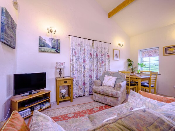 A cozy living room with a flat-screen TV, a floral-patterned armchair, a dining table with chairs, and a large window with a view. - Pickle Cottage (Carnforth)