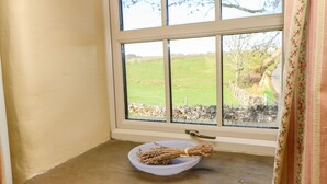 A window with a view of a green field and stone wall, a white plate with twigs on a window sill, and a curtain with a floral pattern. - Cherry Tree Cottage (Kirkby Stephen)
