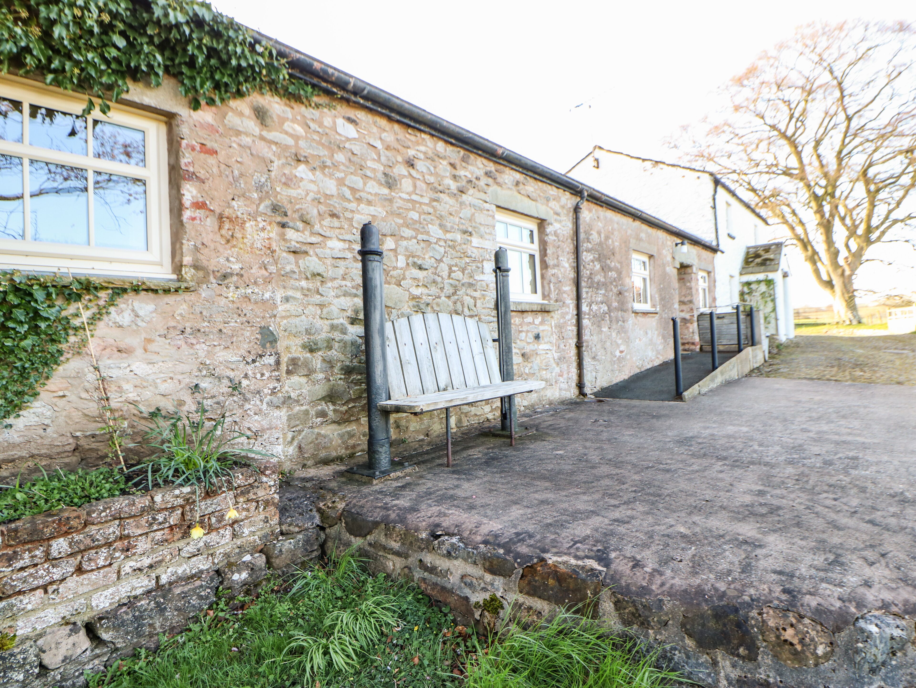 A stone building with a bench outside, a brick wall, and a tree in the background.