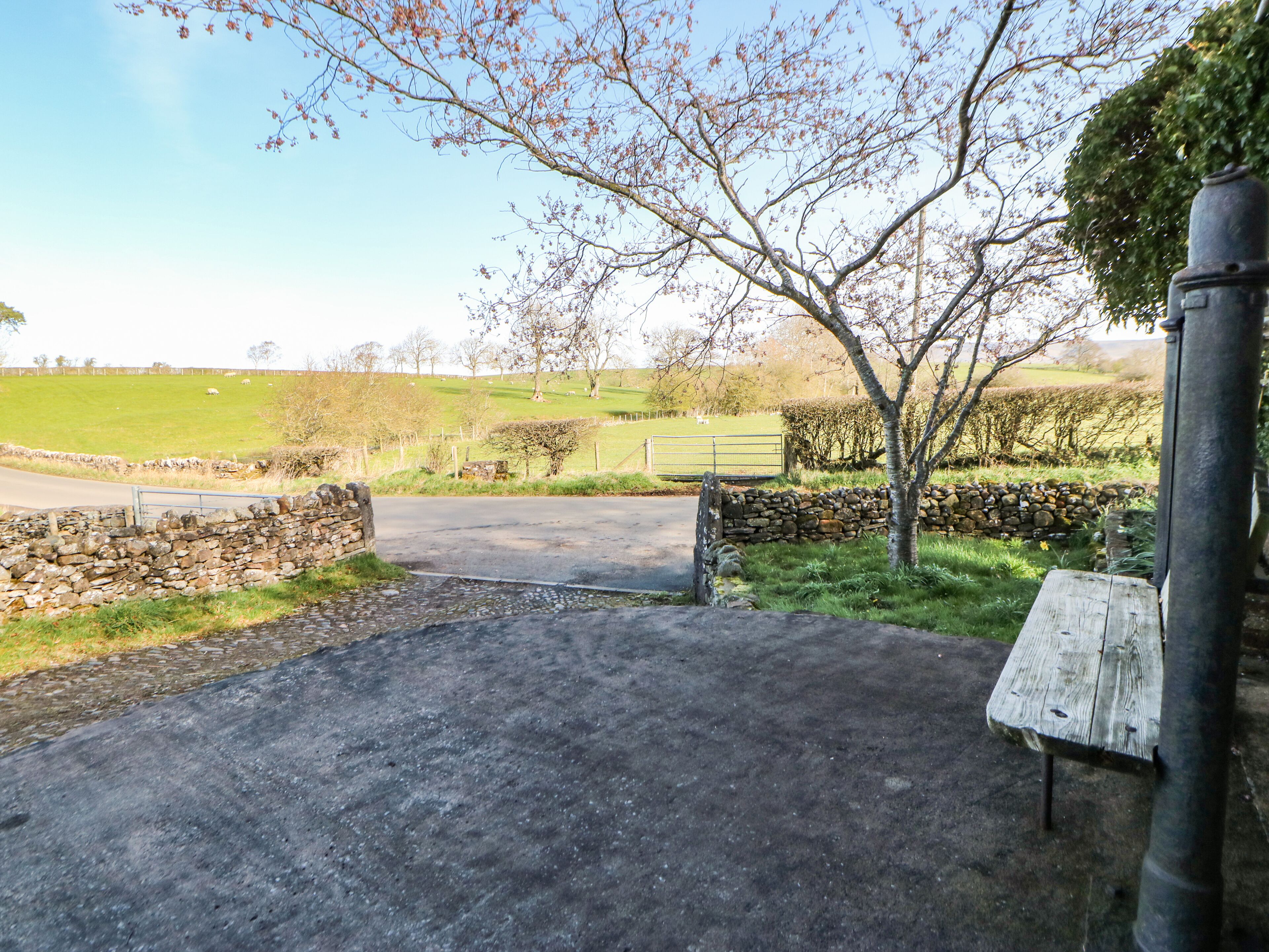 A rural entrance with a stone wall, a wooden bench, and a gate leading to a green field.