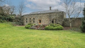 A stone building with a sloped roof, a central door, and a small garden area with shrubs.