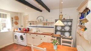 Une cuisine avec une table en bois, des chaises, un lave-linge, un évier et une étagère remplie de vaisselle.