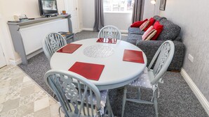 A compact living space with a dining area, a sofa with cushions, a television, and a window with curtains. - Beech Cottage (Tyn-y-Gongl)