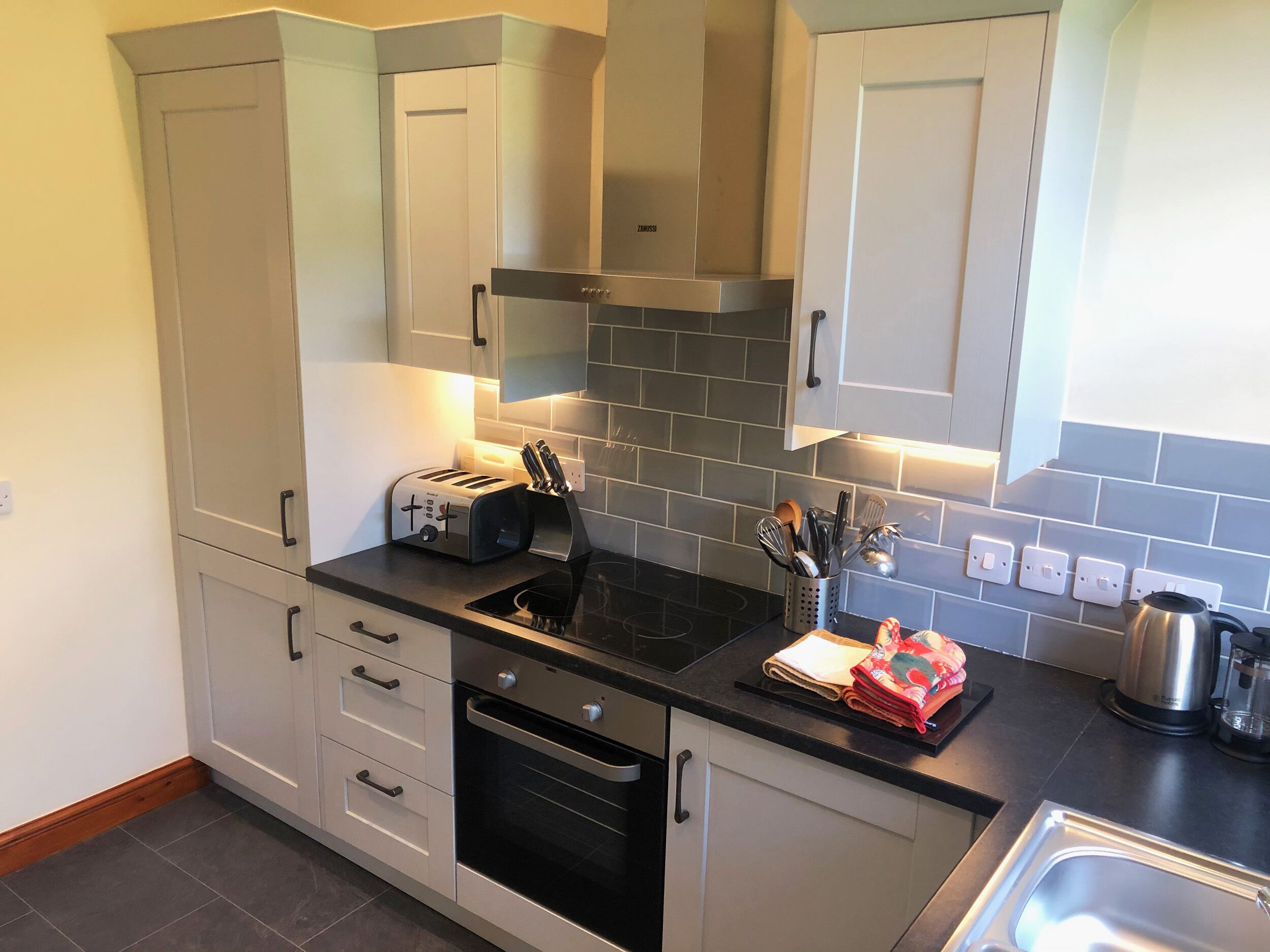 A modern kitchen with white cabinets, a black countertop, a built-in oven, and a stainless steel kettle.